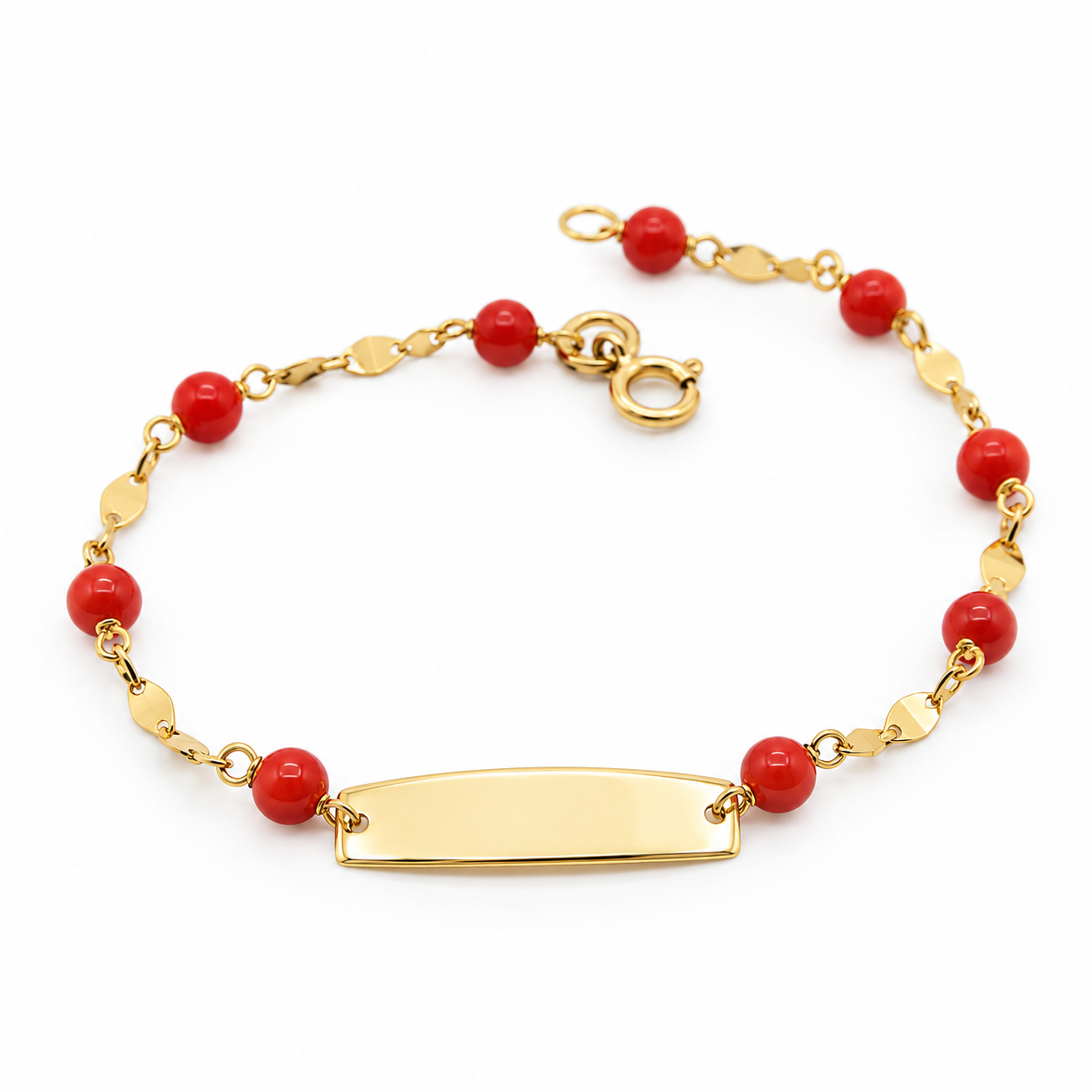 Bracciale da bambino in oro giallo 18 carati con piastrina centrale incisa di dimensioni 16 mm per 5 mm e sferette di corallo rosso alternate lungo la catena, lungo 14,5 cm e dal peso di 0,98 grammi, adagiato su base circolare in marmo con bomboniera e nastro di Battesimo sullo sfondo.